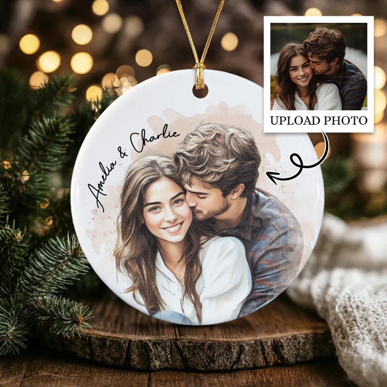 A round white ceramic ornament featuring a custom watercolor-style illustration of a couple. The text "Amelia & Charlie" is printed in black script on the left side. A gold string is attached to the top for hanging purposes.