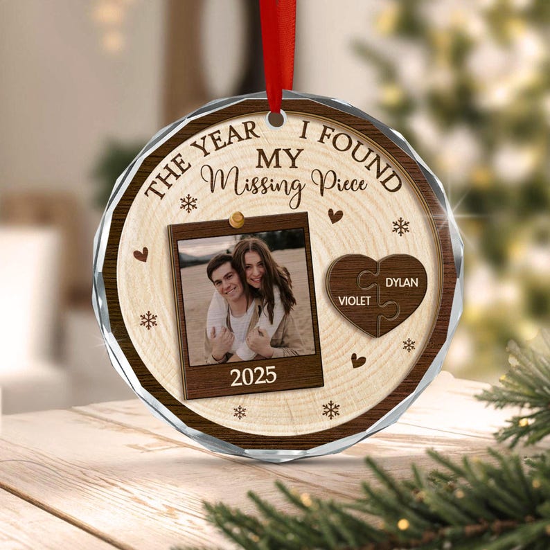 A round, faceted glass ornament with a wood-grain print background. It features the text THE YEAR I FOUND MY Missing Piece, a photo of a couple, the year 2025, and a heart-shaped puzzle piece with the names VIOLET and DYLAN.