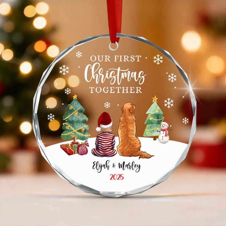 A circular clear glass Christmas ornament with a red ribbon featuring the text OUR FIRST CHRISTMAS TOGETHER, Elijah & Marley, and 2025. The graphic shows a child in striped pajamas and a dog sitting before two trees and a snowman.