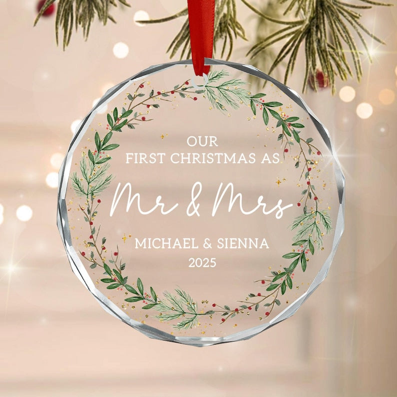 A circular clear acrylic Christmas ornament featuring a red ribbon hanger. The design includes a green pine wreath with red berries and gold flecks, with the text: OUR FIRST CHRISTMAS AS Mr & Mrs MICHAEL & SIENNA 2025.