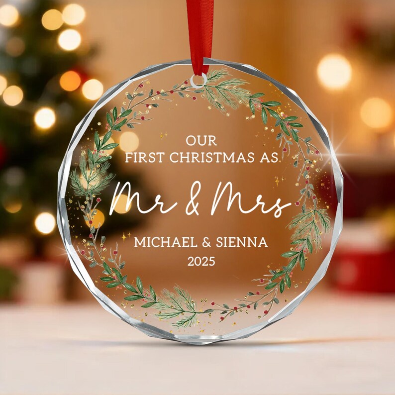 A circular, faceted glass Christmas ornament hanging from a red ribbon. It features a printed wreath of green pine branches and red berries surrounding the text: OUR FIRST CHRISTMAS AS, Mr & Mrs, MICHAEL & SIENNA, 2025.