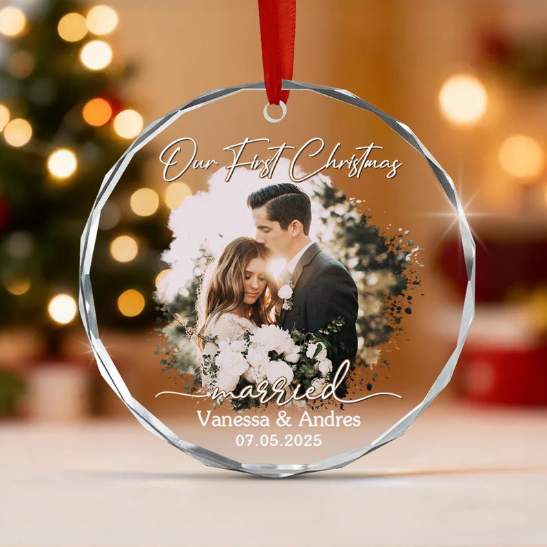 A circular, faceted glass Christmas ornament featuring a printed photograph of a couple, with the text "Our First Christmas", "married", "Vanessa & Andres", and the date "07.05.2025" in white script, suspended by a red ribbon.