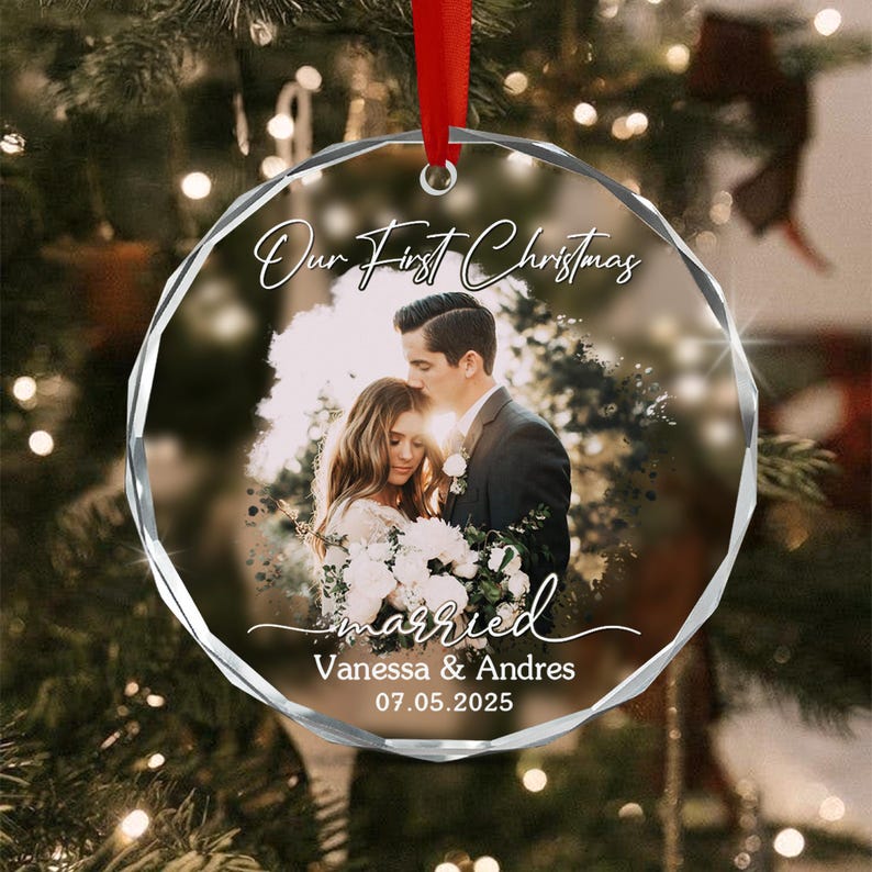 A round, clear acrylic Christmas ornament featuring a printed photograph of a couple in wedding attire. The text reads Our First Christmas, married, Vanessa & Andres, and 07.05.2025. It hangs from a red ribbon.