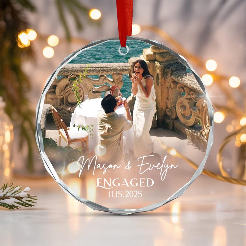A round, faceted glass ornament hanging from a red ribbon. The center features a photograph of a person proposing to another on a terrace overlooking the sea.