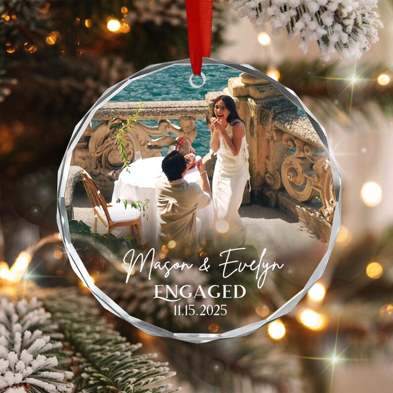 A circular, faceted glass ornament hanging from a red ribbon. The center features a photograph of a couple at a table by the water. White text reads: Mason & Evelyn, ENGAGED, 11.15.2025. The ornament has a clear, reflective edge.