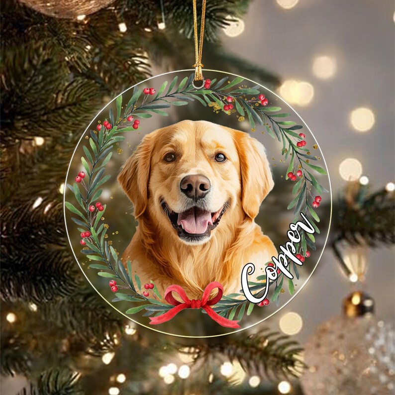 A round, clear acrylic Christmas ornament featuring a portrait of a golden retriever dog centered within a wreath of green leaves and red berries. A red ribbon bow is at the bottom, and the name Copper is written in white script.