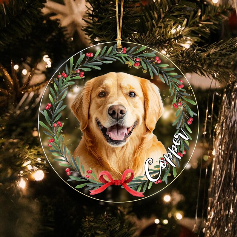 A round Christmas ornament featuring a golden retriever portrait framed by a green wreath with red berries and a red bow at the bottom. The name Copper is written in white cursive script on the right side of the circular ornament.