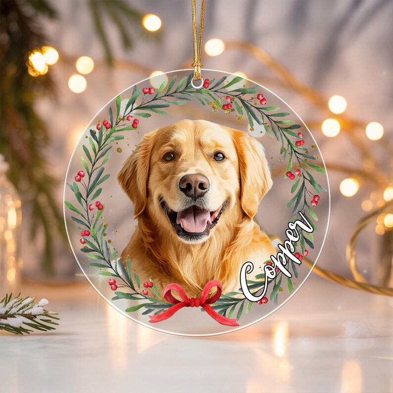 A circular clear acrylic ornament featuring a golden retriever portrait framed by a green leafy wreath with red berries and a red bow at the bottom.