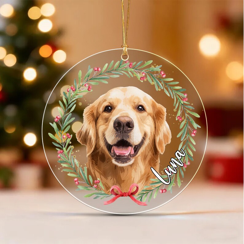 A circular clear acrylic ornament featuring a golden retriever portrait centered within a green wreath with red berries and a red bow at the bottom. The name Luna is printed in white cursive script on the right side of the wreath.