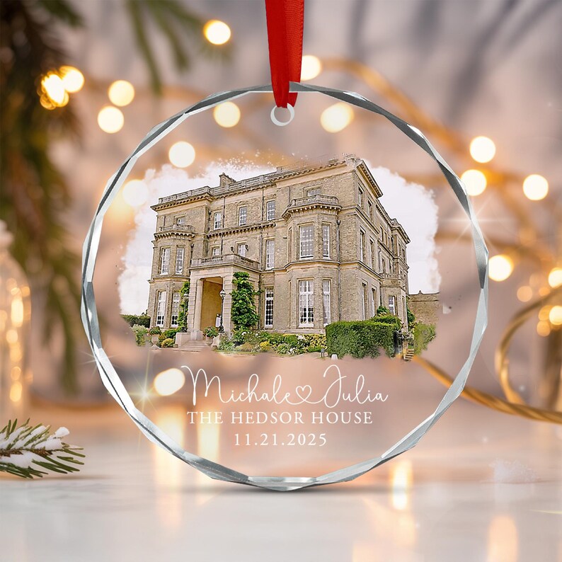 A circular, faceted glass ornament featuring a printed image of a large stone mansion. Below the house, white text reads "Michale & Julia", "THE HEDSOR HOUSE", and "11.21.2025". A red ribbon is attached to the top for hanging.