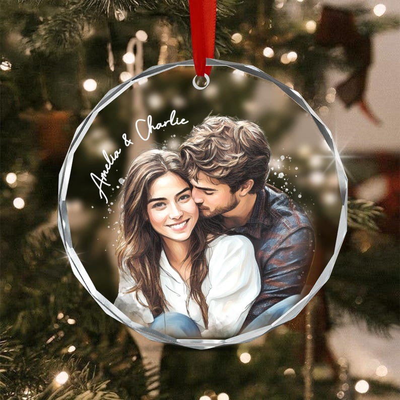A circular clear acrylic ornament featuring a printed illustration of a couple embracing. The woman has long brown hair and wears a white top, while the man has brown hair and a dark shirt. The text Amelia & Charlie is printed.