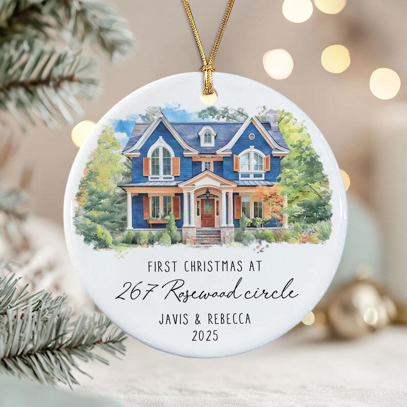 A round white ceramic ornament featuring a watercolor illustration of a blue two-story house with brown shutters. The text reads: FIRST CHRISTMAS AT 267 Rosewood circle JAVIS & REBECCA 2025.