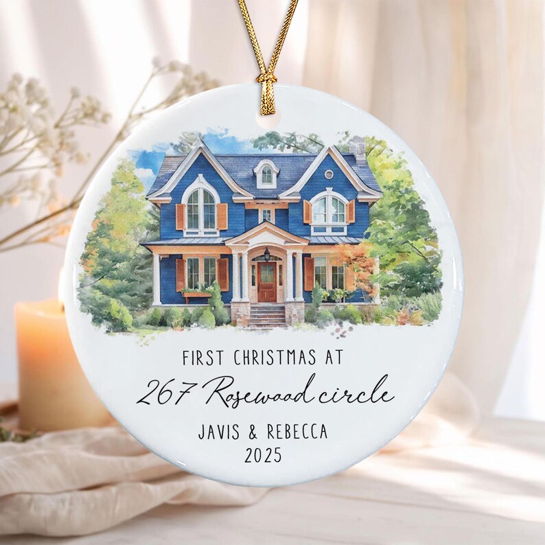 A round white ceramic ornament featuring a watercolor illustration of a blue two-story house with orange shutters. The text reads: FIRST CHRISTMAS AT 267 Rosewood circle JAVIS & REBECCA 2025. It hangs from a gold cord.