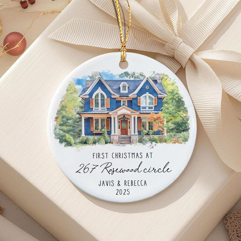 A round white ceramic ornament featuring a watercolor illustration of a blue two-story house with brown shutters. The text reads: FIRST CHRISTMAS AT 267 Rosewood circle JAVIS & REBECCA 2025.