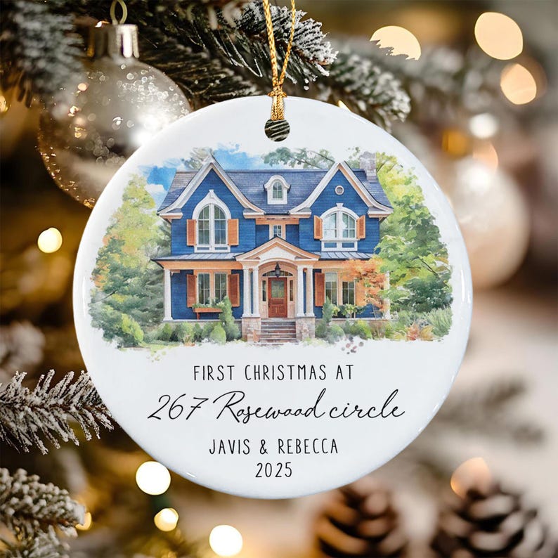 A round white ceramic ornament featuring a watercolor illustration of a blue two-story house with brown shutters. The text reads: FIRST CHRISTMAS AT 267 Rosewood circle JAVIS & REBECCA 2025.