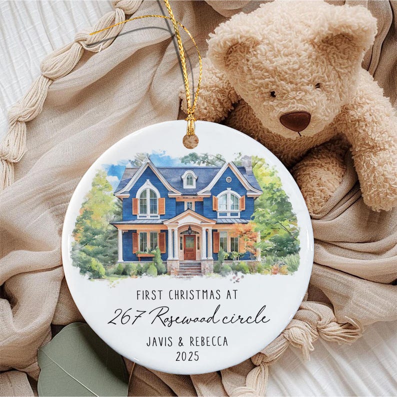 A round white ceramic ornament featuring a watercolor illustration of a blue two-story house with orange shutters. The text reads: FIRST CHRISTMAS AT 267 Rosewood circle JAVIS & REBECCA 2025.