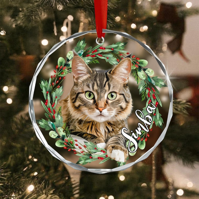 A round, clear, faceted glass ornament hanging from a red ribbon. It features a printed image of a brown tabby cat centered within a wreath of green pine needles and red berries, with the name Simba written in white script text.