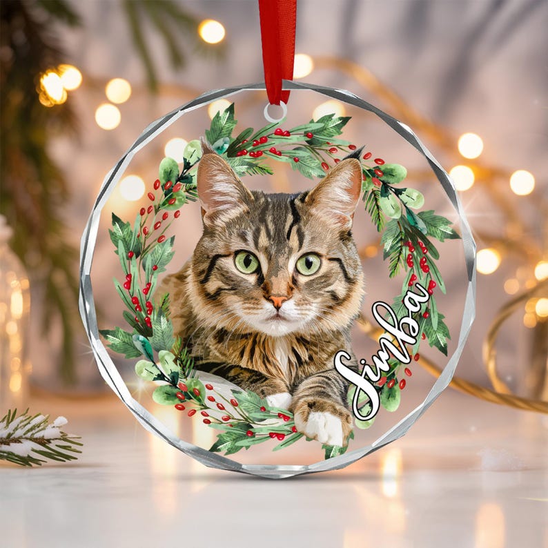 A circular, faceted glass Christmas ornament featuring a brown tabby cat centered within a wreath of green holly leaves and red berries. The name Simba is written in white cursive script on the right side.