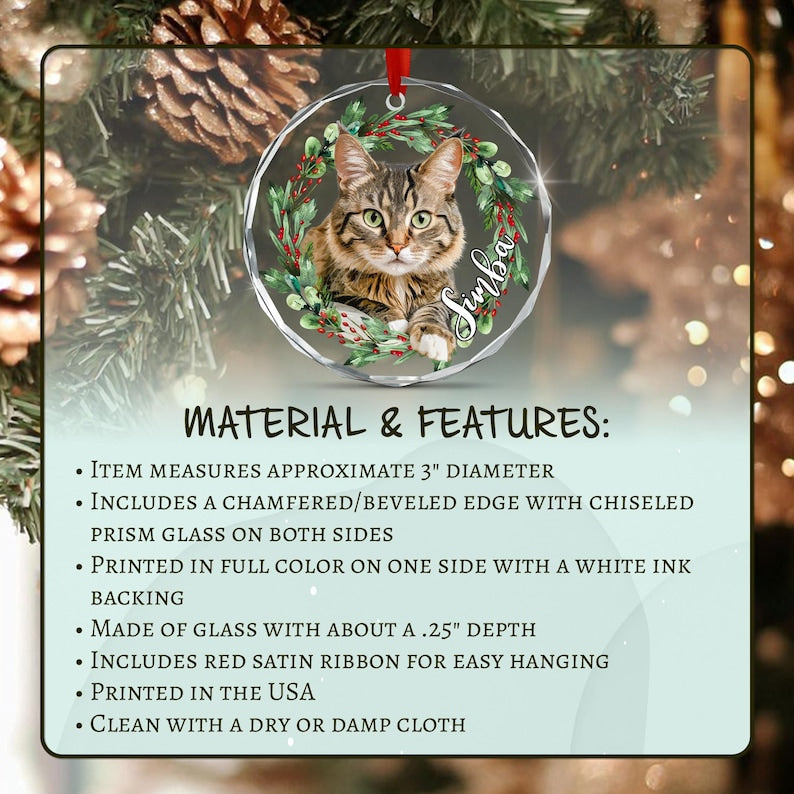 A circular glass ornament featuring a brown tabby cat inside a green wreath with red berries and the name Simba in white script. It has a beveled edge, a red satin ribbon, and measures 3 inches in diameter with a 0.25 inch depth.