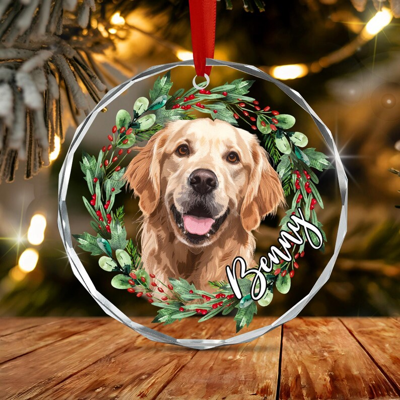A round, faceted glass Christmas ornament featuring a golden retriever portrait framed by a wreath of green leaves and red berries. The name Benny is written in white cursive script on the right side.