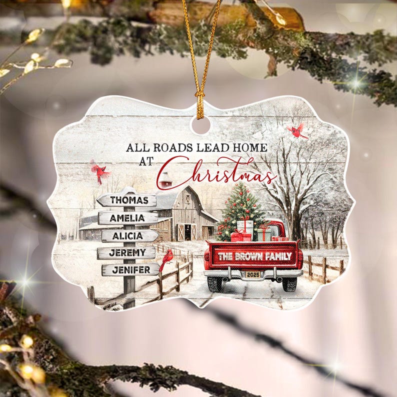 A decorative ornament featuring a winter scene with a red pickup truck, a barn, and a signpost with the names Thomas, Amelia, Alicia, Jeremy, and Jenifer. The text reads All Roads Lead Home At Christmas and The Brown Family 2025.