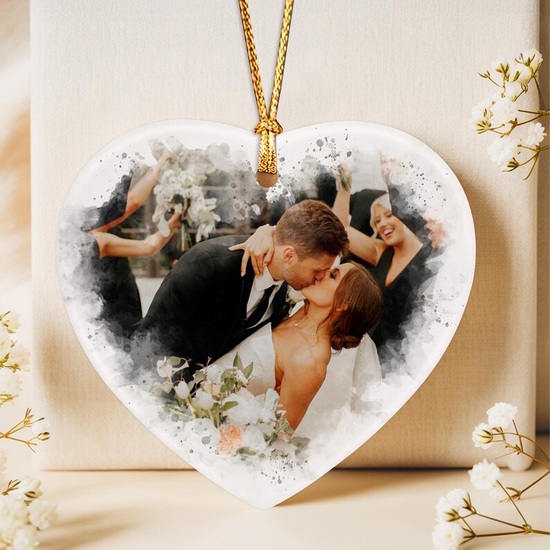 A heart-shaped ceramic ornament featuring a watercolor-style print of a couple kissing at a wedding. The image shows a person in a black suit and a person in a white dress holding a bouquet, with guests in the background.