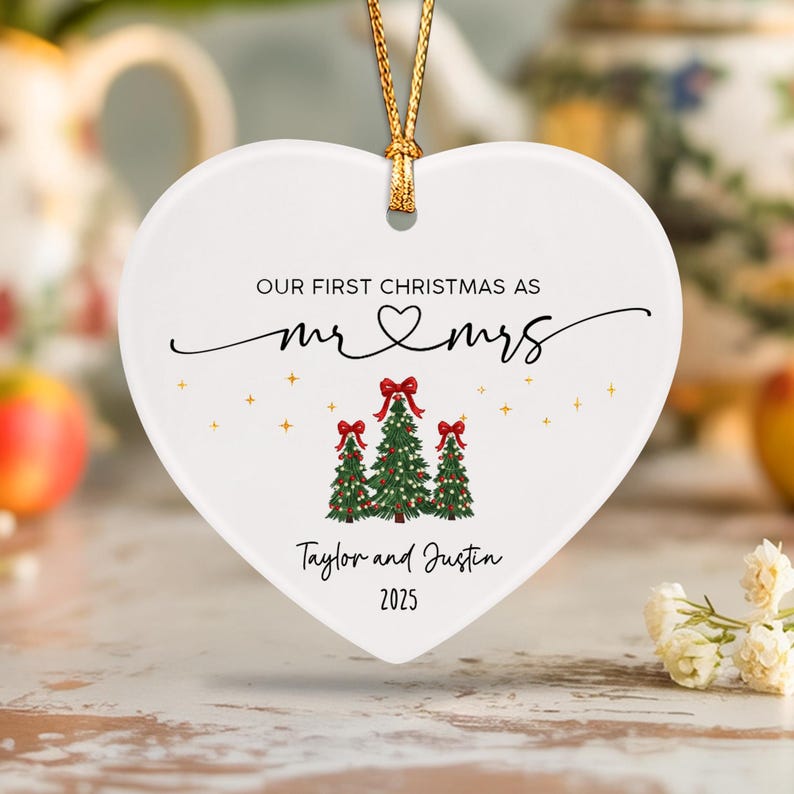 A white heart-shaped ceramic ornament hanging from a gold string. It features the text "OUR FIRST CHRISTMAS AS mr & mrs", "Taylor and Justin", and "2025".