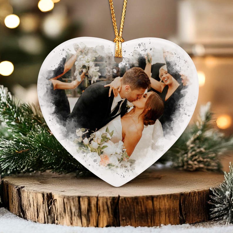 A heart-shaped ceramic ornament featuring a watercolor-style print of a couple kissing at a wedding. The image shows a person in a black suit and a person in a white dress holding a bouquet. A gold cord is attached for hanging.