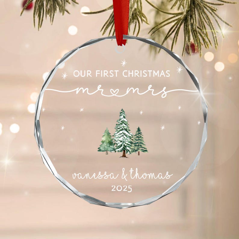 A circular clear glass Christmas ornament with faceted edges, featuring a red ribbon hanger. The text reads OUR FIRST CHRISTMAS, mr & mrs, vanessa & thomas, and 2025. A watercolor illustration of three pine trees is centered.