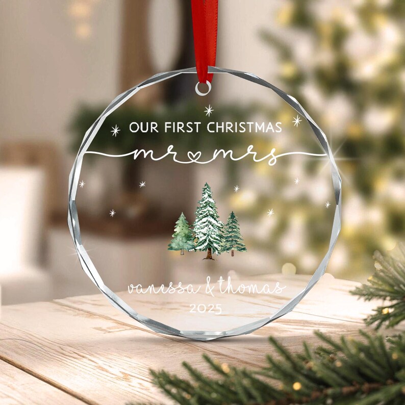 A circular, faceted glass Christmas ornament hanging from a red ribbon. It features the text "OUR FIRST CHRISTMAS mr & mrs", "vanessa & thomas", and "2025" in white, along with a watercolor illustration of three green pine trees.