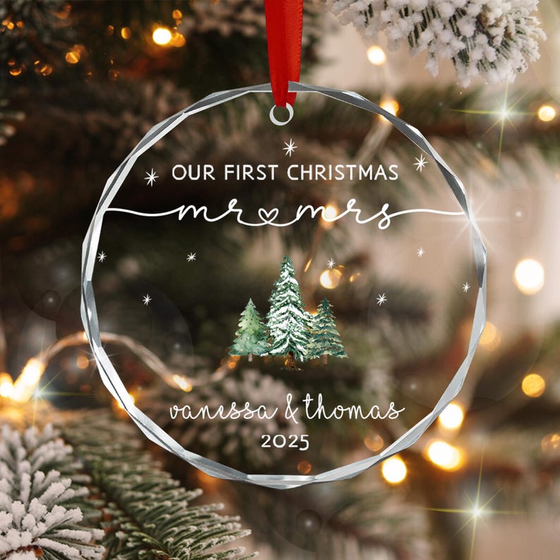 A circular clear acrylic ornament with faceted edges, featuring a red ribbon hanger. The text reads OUR FIRST CHRISTMAS, mr & mrs, vanessa & thomas, and 2025. A watercolor illustration of three evergreen trees sits in the center.