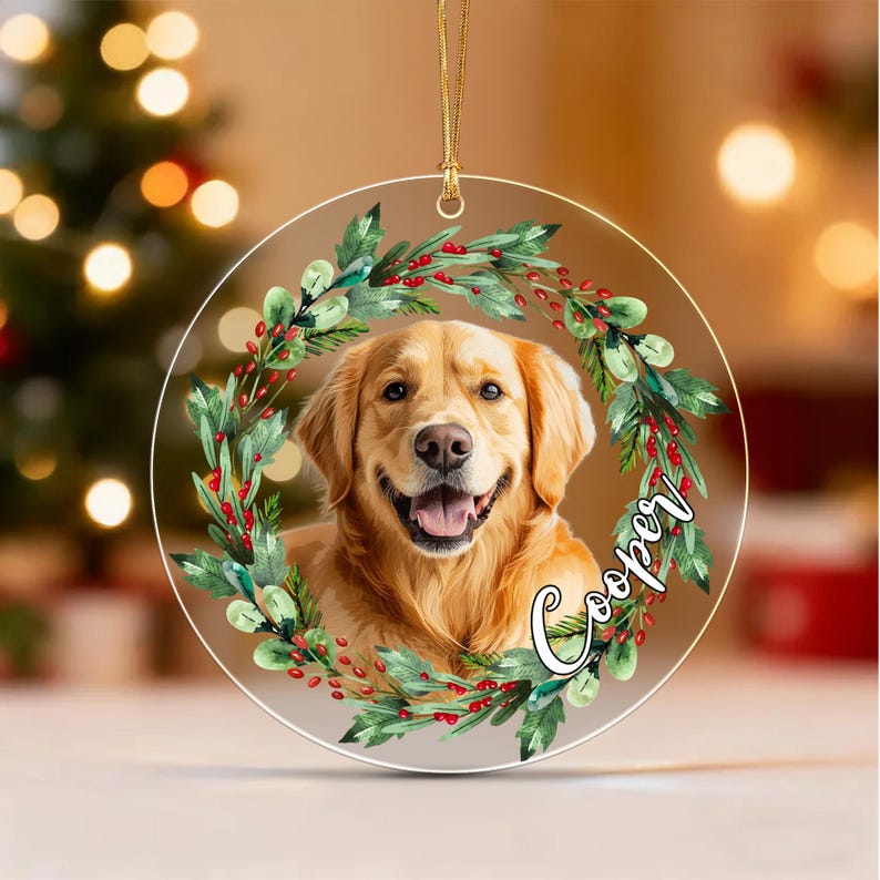 A circular clear acrylic ornament featuring a golden retriever portrait framed by a watercolor wreath of green leaves and red berries. The name Cooper is printed in white cursive script on the right side, suspended by a gold cord.