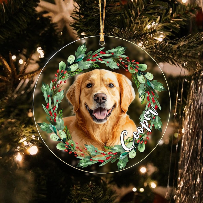 A round, flat, transparent ornament featuring a golden retriever portrait framed by a green wreath with red berries. The name Cooper is printed in white script on the right side. A gold string is attached to the top for hanging.