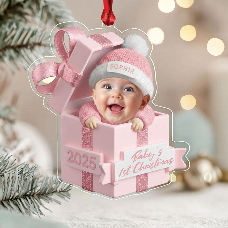 A clear acrylic Christmas ornament shaped like a pink gift box with a baby wearing a pink knit hat with the name SOPHIA printed on it. The box features the text 2025 and Baby's 1st Christmas on a white banner.