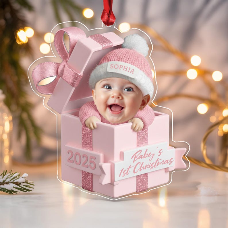 A clear acrylic Christmas ornament shaped like a pink gift box with a baby wearing a pink knit hat with a white pom-pom. The hat features the name SOPHIA. The box displays the text 2025 and Baby's 1st Christmas on a banner.