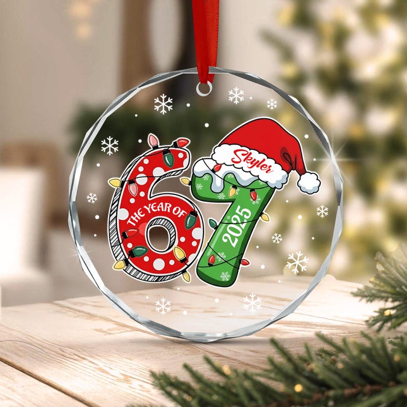 A circular clear glass ornament with a red ribbon hanger. The design features the number 67, with the 6 in red with white polka dots and the 7 in green. The 7 wears a Santa hat with the name Skyler and the year 2025 printed on it.