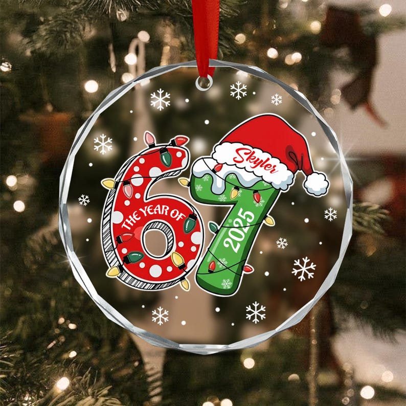 A round, clear, faceted glass Christmas ornament hanging from a red ribbon. The design features the number 67 with red polka dots and green coloring, wrapped in string lights, with the text THE YEAR OF, 2025, and Skyler.