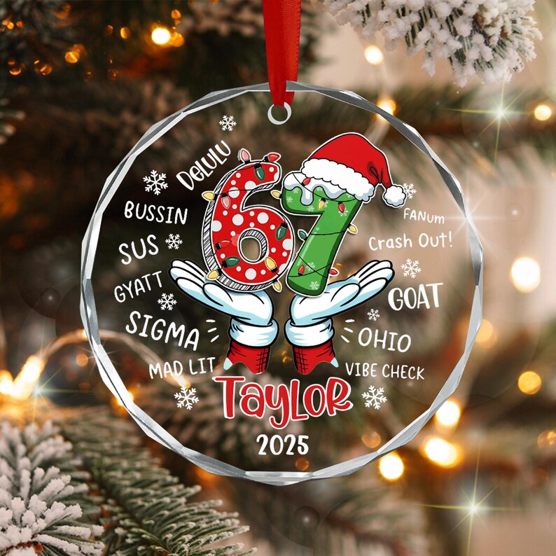 A circular clear acrylic Christmas ornament featuring the number 67 in red and green with a Santa hat, held by cartoon gloved hands. It includes the name Taylor, the year 2025, and slang terms like Delulu, Bussin, Sus, and Goat.