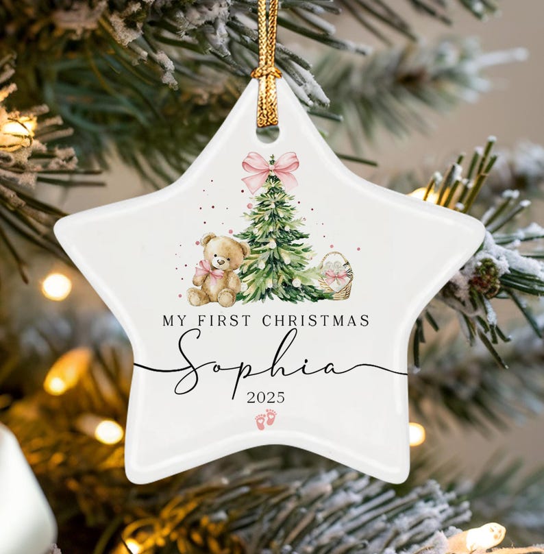 A white star-shaped ceramic ornament featuring a watercolor illustration of a teddy bear, a Christmas tree, and a pink bow. The text reads "MY FIRST CHRISTMAS", "Sophia", and "2025" with small pink baby footprint icons below.