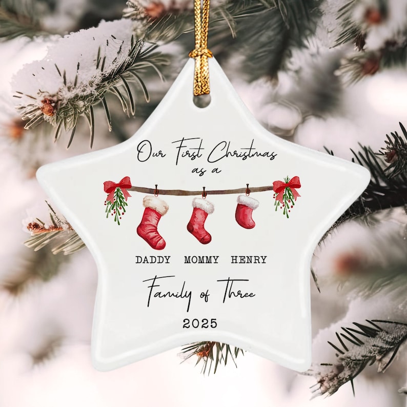 A white star-shaped ceramic ornament with a gold hanging cord. It features a printed illustration of three red stockings hanging from a branch with mistletoe and red bows. Text reads: Our First Christmas as a Family of Three 2025.