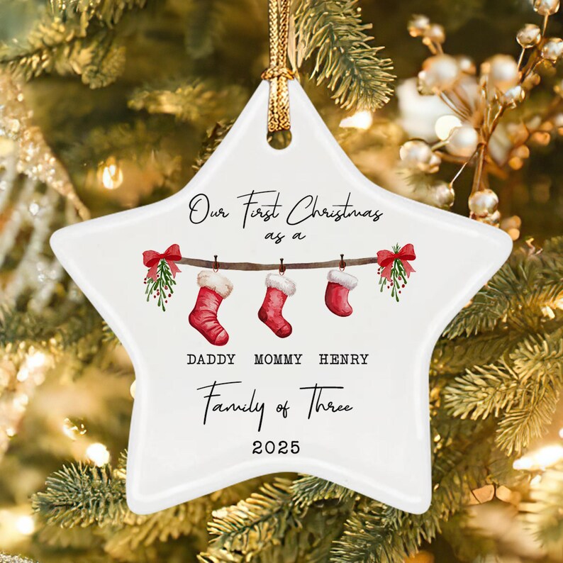 A white star-shaped ceramic ornament featuring a printed illustration of three red Christmas stockings hanging from a branch with mistletoe. Text reads: Our First Christmas as a Family of Three, DADDY MOMMY HENRY, and 2025.