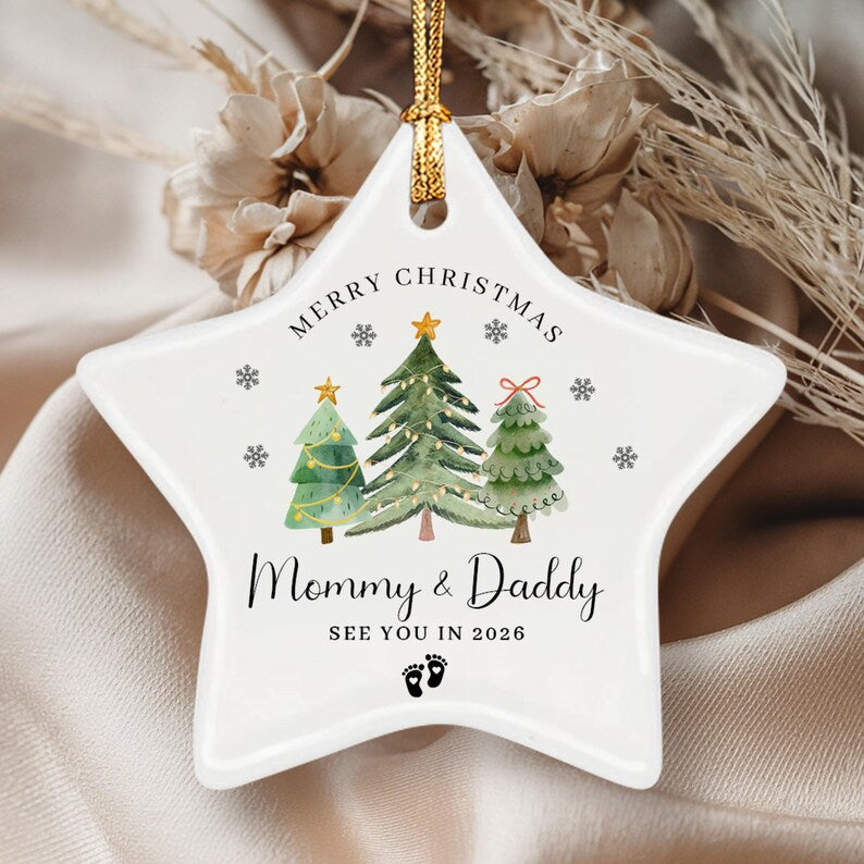 A white star-shaped ceramic ornament featuring a watercolor illustration of three Christmas trees, snowflakes, and the text Merry Christmas, Mommy & Daddy, SEE YOU IN 2026, and a pair of small black baby footprints.