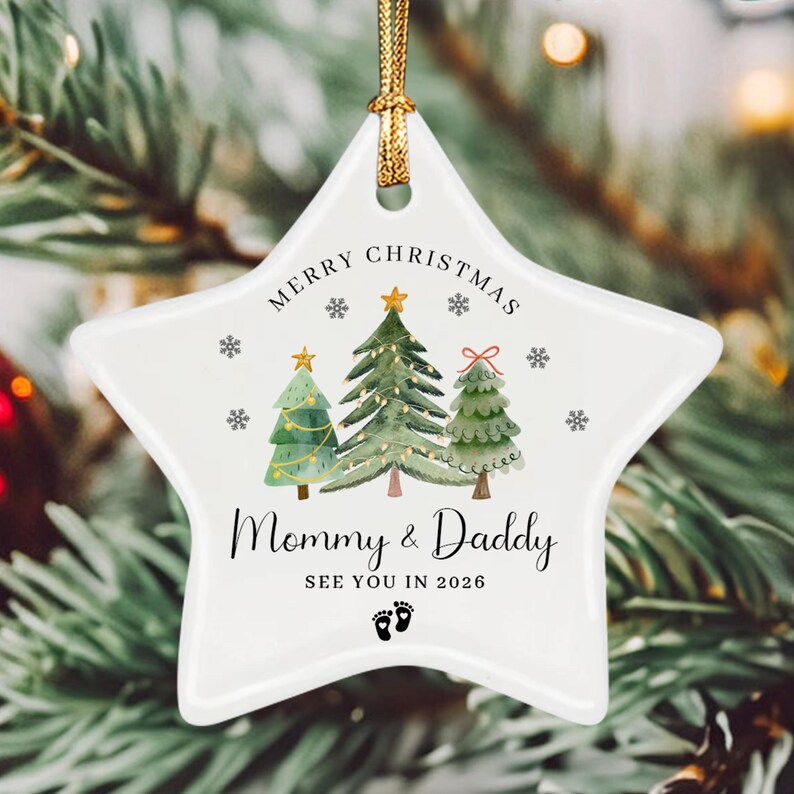 A white star-shaped ceramic ornament featuring a watercolor illustration of three Christmas trees, snowflakes, and the text Merry Christmas, Mommy & Daddy, SEE YOU IN 2026, and a pair of baby footprints with a gold hanging cord.
