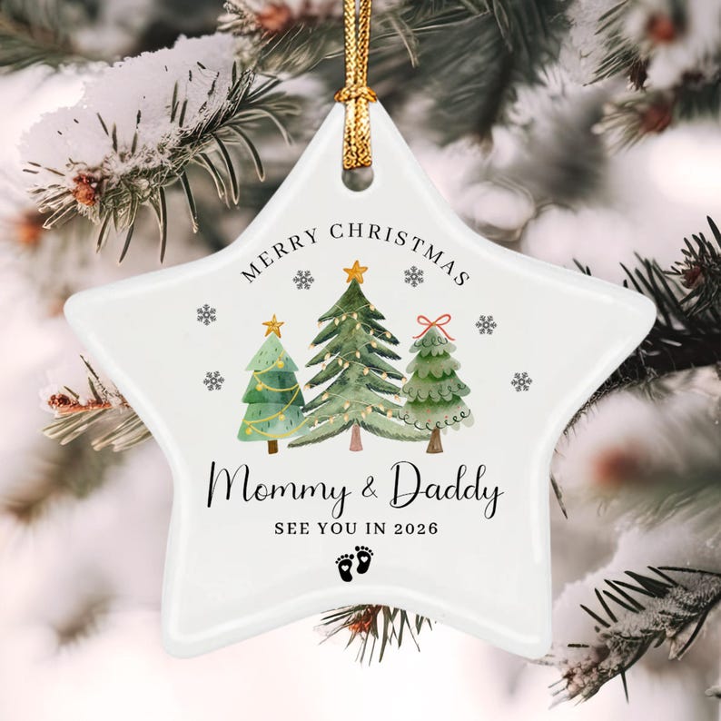 A white star-shaped ceramic ornament featuring a watercolor illustration of three Christmas trees, snowflakes, and the text Merry Christmas, Mommy & Daddy, SEE YOU IN 2026, and a pair of baby footprints, suspended by a gold cord.