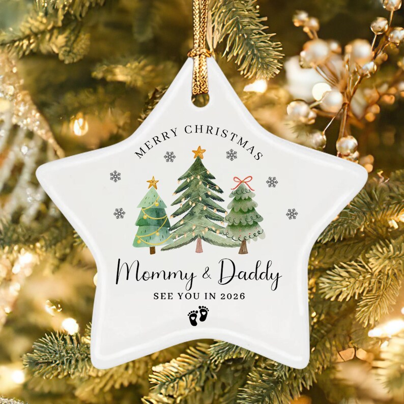 A white star-shaped ceramic Christmas ornament featuring three watercolor-style pine trees, small snowflakes, and the text Merry Christmas, Mommy & Daddy, SEE YOU IN 2026, and a pair of small black baby footprints at the bottom.