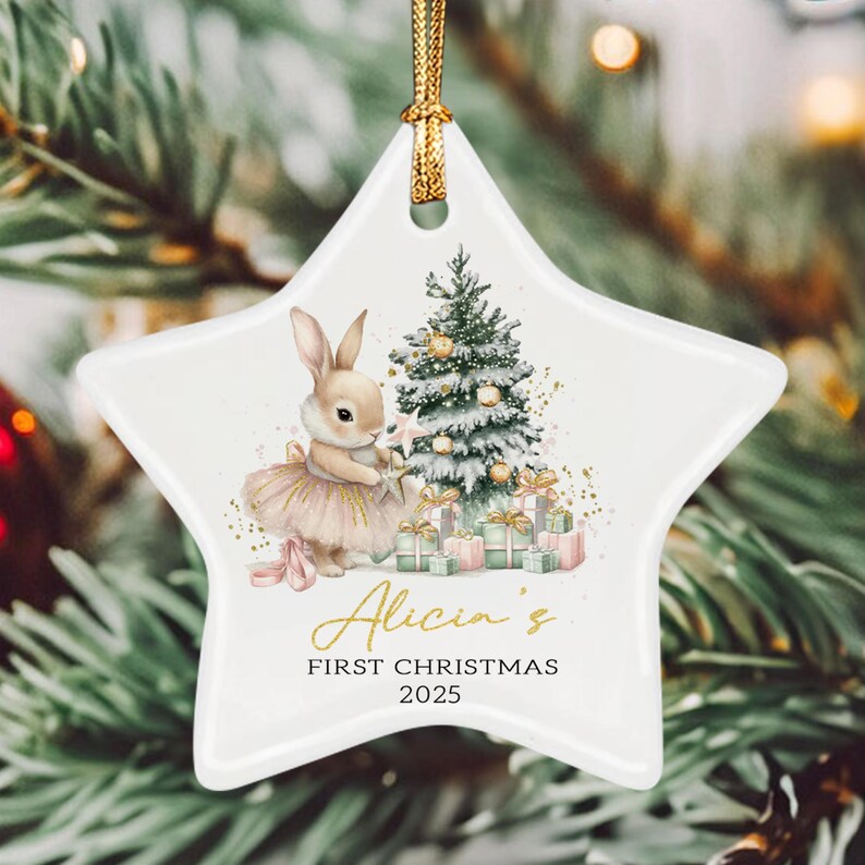 A white star-shaped ceramic ornament featuring a watercolor illustration of a rabbit in a tutu next to a decorated Christmas tree and gifts. The text reads Alicia's FIRST CHRISTMAS 2025.