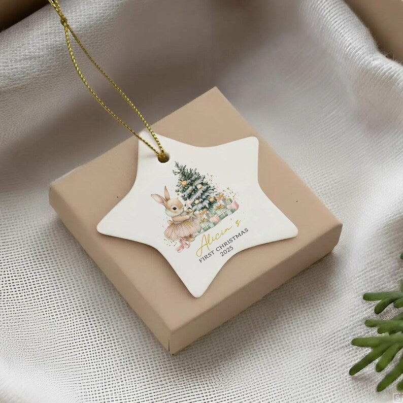 A star-shaped white ceramic ornament featuring a watercolor illustration of a rabbit wearing a tutu next to a decorated Christmas tree. The text reads Alicia's FIRST CHRISTMAS 2025 in gold and black script.