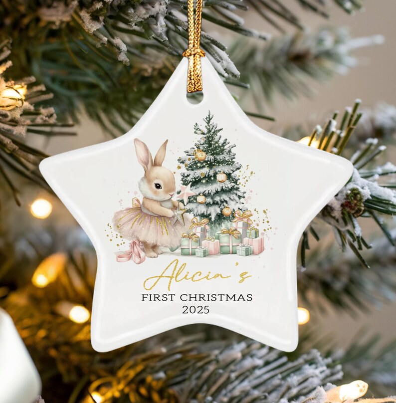 A white star-shaped ceramic ornament featuring a watercolor illustration of a bunny in a tutu next to a decorated Christmas tree and gifts. The text reads Alicia's FIRST CHRISTMAS 2025 in gold and black script and block lettering.