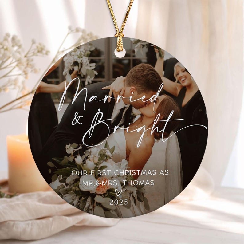 A round ceramic Christmas ornament featuring a photograph of a couple kissing at their wedding. The text reads Married & Bright, OUR FIRST CHRISTMAS AS MR. & MRS. THOMAS, and 2025. It hangs from a gold cord.