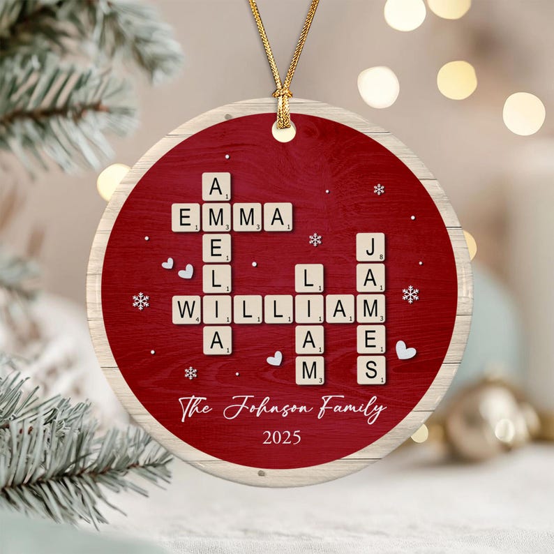 A round ceramic ornament with a red wood-grain background featuring a crossword-style arrangement of letter tiles spelling AMELIA, WILLIAM, and JAMES. Below the tiles, the text reads The Johnson Family and the year 2025.